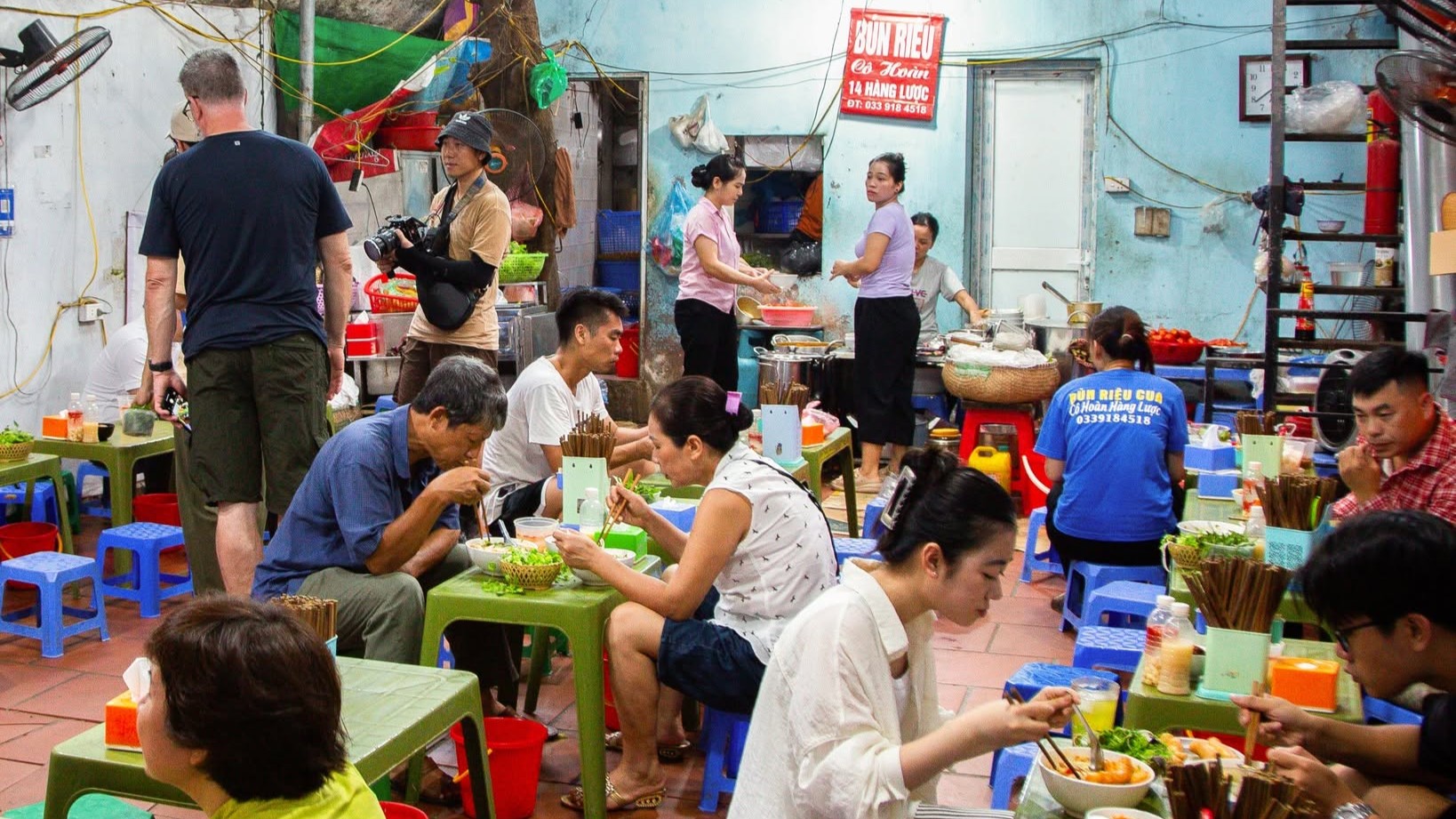 Diners eating together on low stools at a busy Vietnamese street food restaurant.